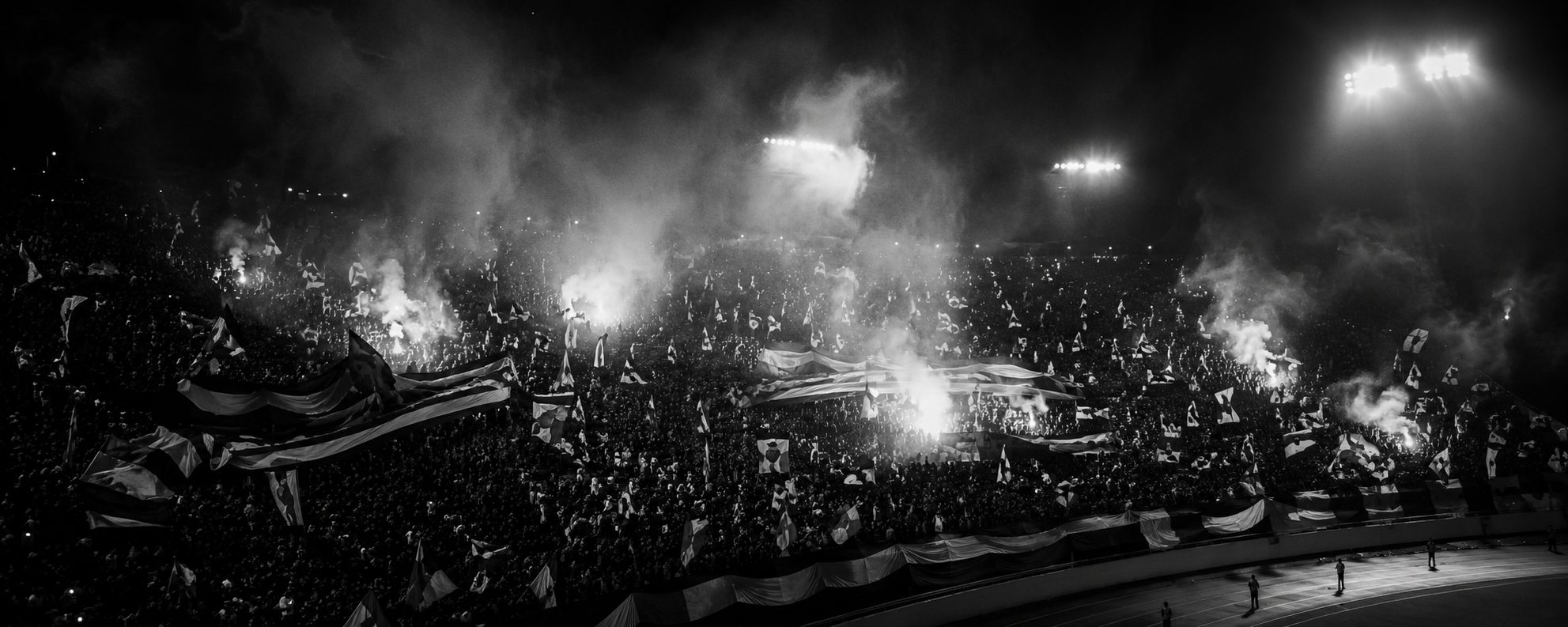 Arquibancada lotada à noite, bandeiras e fumaça sob os refletores